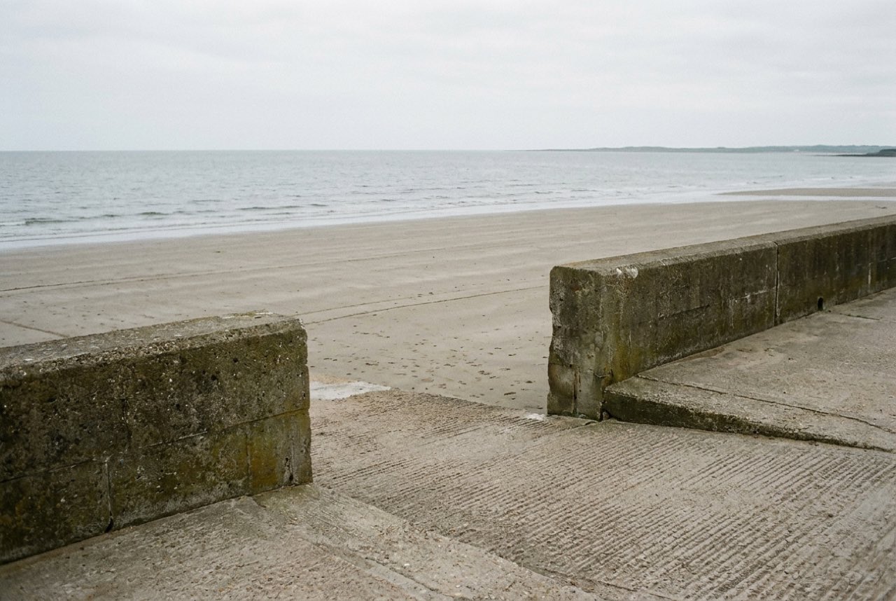 A level gap in a low sea wall with a flat concrete ramp running down onto wide firm sand and calm water, even grey light
