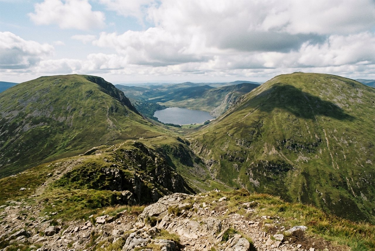 A saddle between two rounded green summits with a lake far below on the far side, cloud shadow moving across