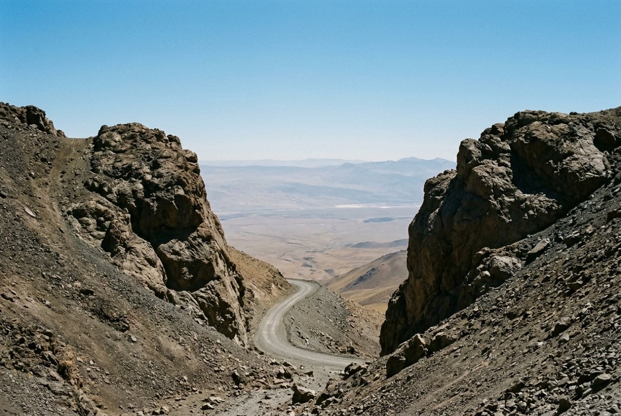 A high mountain pass with the road dropping away into a hazy plain beyond, bare rock either side, thin clear air