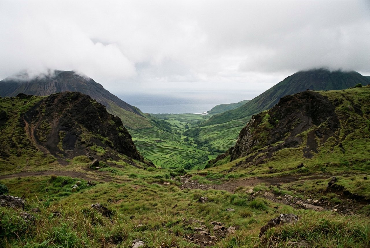 A saddle between two volcanic cones opening onto a green valley of rice terraces running down to the sea, wet season cloud