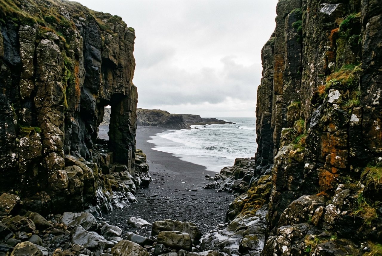 A gap in a basalt sea cliff opening onto grey Atlantic water and a black beach, moss on the rock, low bright cloud