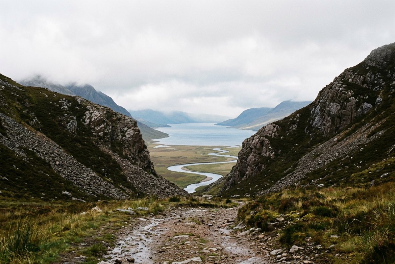 A col between two bare quartzite hills opening onto a wide empty Scottish strath with a river and a sea loch beyond, low cloud