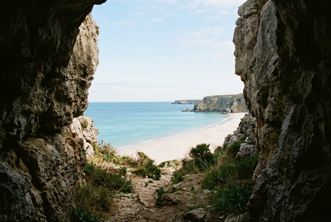 A narrow gap in a line of coastal cliffs opening onto a pale crescent beach and calm turquoise water beyond, clear morning light